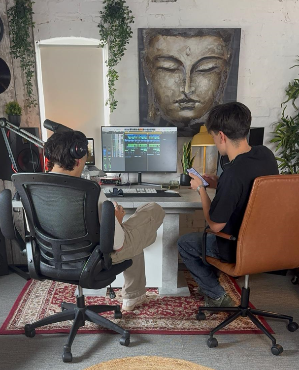 Two people working together at the desk, seen from behind, with a Buddha painting and hanging plants on the wall above the monitors.